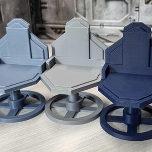 May include: Three miniature chairs in shades of blue and gray. Each chair has a geometric design with a backrest, seat, and a circular base. The chairs are likely for a model or display.
