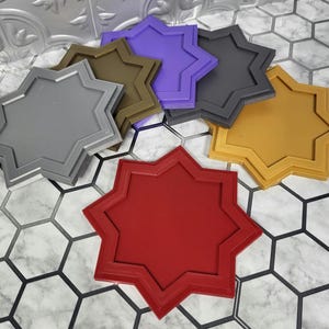 May include: A collection of eight-pointed star-shaped coasters in various colors, including red, gold, gray, olive green, purple, and black. The coasters are made of a solid material and have a raised edge.