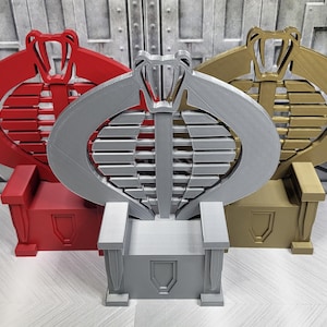 May include: Three Cobra Commander throne replicas in red, silver, and gold. Each throne features a cobra emblem backrest and a rectangular base with a shield design. The thrones are displayed on a light-colored surface.