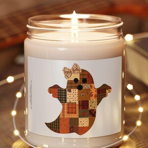 May include: A clear glass candle jar with a white wax fill and a lit wick, sitting on a round wooden base. The label features a ghost graphic composed of various brown, orange, and tan patchwork fabric patterns with a small plaid bow on top.