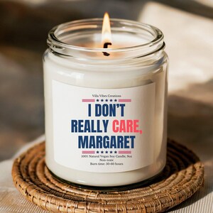 May include: A white soy candle in a glass jar with a red, white, and blue label that reads "I don't really care, Margaret." The candle is on a woven coaster.