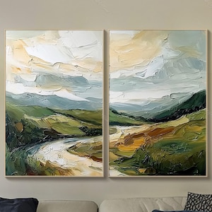 May include: Two framed landscape paintings with textured brushstrokes. The artwork depicts a river winding through green hills under a cloudy sky. The paintings are displayed above a beige sofa with blue cushions.
