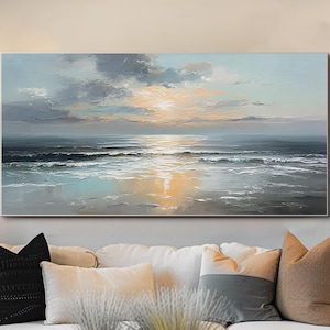 Op de afbeelding: Een groot rechthoekig canvas schilderij dat een zeegezicht met zonsondergang uitbeeldt. Het kunstwerk toont een mix van blauw, grijs en gouden tinten, die de oceaangolven en de weerspiegeling van de lucht vastleggen. Het schilderij hangt boven een witte bank.