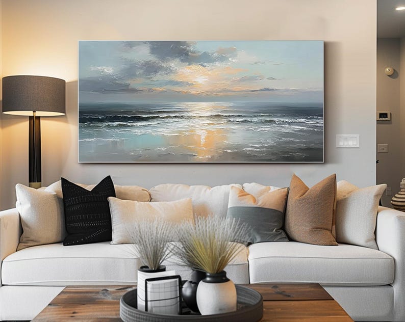 Op de afbeelding: Een groot rechthoekig canvas schilderij dat een zeegezicht met zonsondergang uitbeeldt. Het kunstwerk toont een mix van blauw, grijs en gouden tinten, die de oceaangolven en de weerspiegeling van de lucht vastleggen. Het schilderij hangt boven een witte bank.