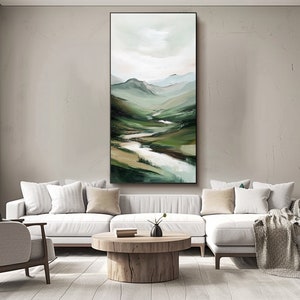 May include: Vertical landscape painting depicting a valley with a river winding through green hills. The artwork features a palette of greens, browns, and whites, with a cloudy sky. The painting is framed in black and hangs above a white sofa.