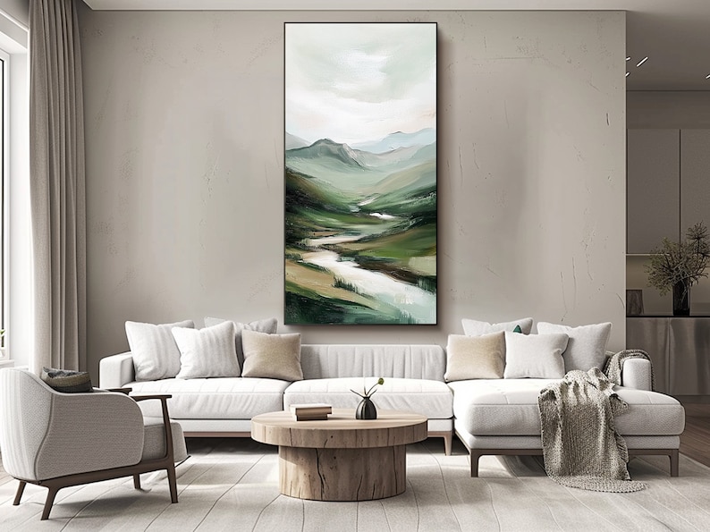 May include: Vertical landscape painting depicting a valley with a river winding through green hills. The artwork features a palette of greens, browns, and whites, with a cloudy sky. The painting is framed in black and hangs above a white sofa.