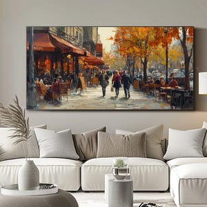 May include: A painting of a Parisian street scene with people walking and sitting at outdoor cafes. The buildings are painted in warm tones, and the trees are in autumn colors. The painting is framed in a black frame.