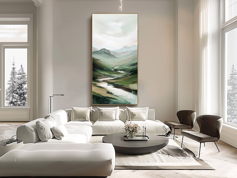 May include: An abstract landscape painting with a gold frame. The artwork depicts a valley with a river, rendered in shades of green, brown, and white. The painting hangs above a white sectional sofa in a modern living room.