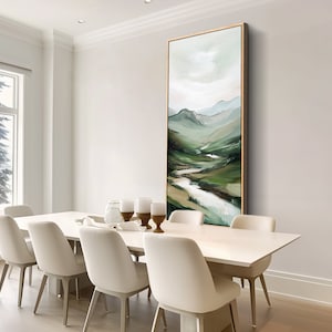May include: An abstract landscape painting in a vertical rectangular format. The artwork features a river winding through green hills and mountains, with a palette of greens, blues, and whites. The painting is framed in a gold tone and hangs in a dining room.