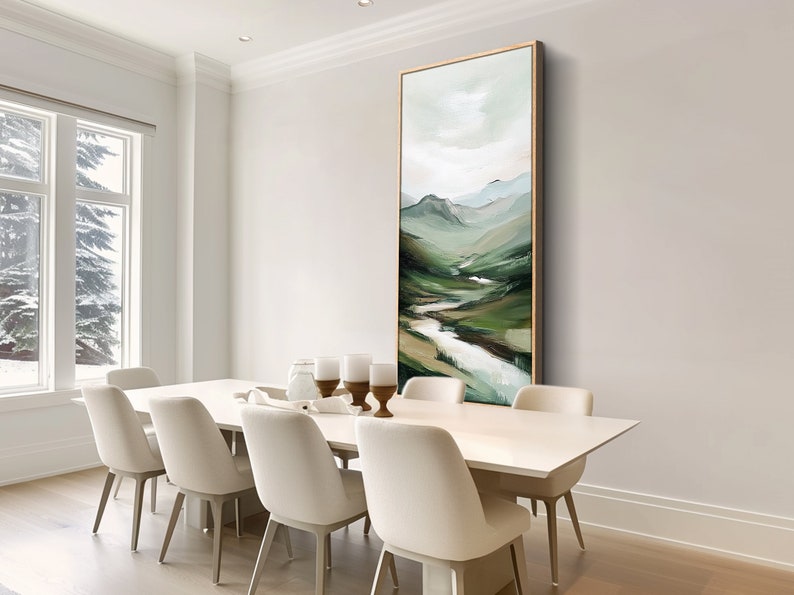 May include: An abstract landscape painting in a vertical rectangular format. The artwork features a river winding through green hills and mountains, with a palette of greens, blues, and whites. The painting is framed in a gold tone and hangs in a dining room.