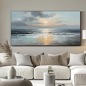 Op de afbeelding: Grote rechthoekige canvas kunst die een zeegezicht bij zonsondergang uitbeeldt. Het schilderij toont een mix van blauw, grijs en oranje, met zichtbare penseelstreken. Het kunstwerk is zwart omlijst en hangt boven een witte bank.