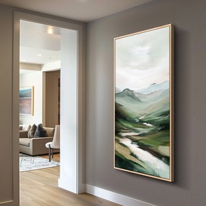May include: A framed landscape painting depicts a valley with rolling green hills and a winding river. The artwork features a palette of greens, whites, and blues, with a light-colored sky. The painting is displayed in a room with a neutral-colored wall.