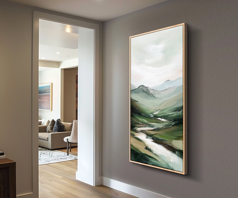 May include: A framed landscape painting depicts a valley with rolling green hills and a winding river. The artwork features a palette of greens, whites, and blues, with a light-colored sky. The painting is displayed in a room with a neutral-colored wall.