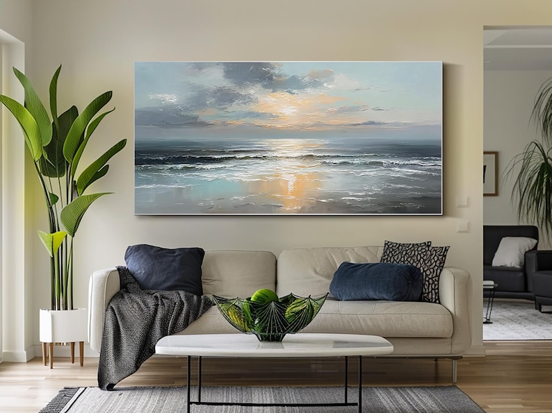 Op de afbeelding: Olieverfschilderij van een zeegezicht met zonsondergang. De lucht is gevuld met grijze en witte wolken en de zon gaat onder boven de oceaan. De golven slaan op de kust en het water weerspiegelt de kleuren van de lucht. Het schilderij hangt boven een beige bank.