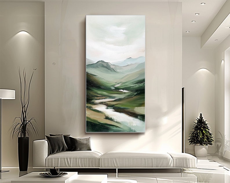 May include: Vertical landscape painting depicting a serene valley with rolling green hills, a winding river, and a soft, cloudy sky. The artwork uses a palette of greens, blues, and whites, creating a calming and natural aesthetic. The painting is displayed in a modern living room setting.