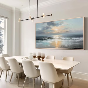 Op de afbeelding: Een olieverfschilderij van een zeegezicht hangt boven een witte eettafel met acht stoelen. Het kunstwerk toont een zonsondergang boven de oceaan, met tinten blauw, grijs en goud. Boven de tafel hangt een moderne kroonluchter.