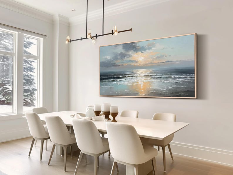 Op de afbeelding: Een olieverfschilderij van een zeegezicht hangt boven een witte eettafel met acht stoelen. Het kunstwerk toont een zonsondergang boven de oceaan, met tinten blauw, grijs en goud. Boven de tafel hangt een moderne kroonluchter.