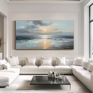 Op de afbeelding: Groot rechthoekig olieverfschilderij dat een zeegezicht met zonsondergang uitbeeldt. Het kunstwerk heeft een palet van blauw, grijs en geel, met de zon die reflecteert op het water. Het schilderij hangt boven een witte hoekbank in een moderne woonkamer.