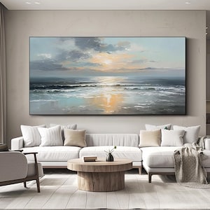 Op de afbeelding: Groot rechthoekig olieverfschilderij dat een zeegezicht met zonsondergang uitbeeldt. Het kunstwerk toont tinten blauw, grijs en goud, met zichtbare penseelstreken. Het schilderij hangt boven een witte hoekbank in een moderne woonkamer.