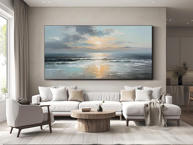 Op de afbeelding: Groot rechthoekig olieverfschilderij dat een zeegezicht met zonsondergang uitbeeldt. Het kunstwerk toont tinten blauw, grijs en goud, met zichtbare penseelstreken. Het schilderij hangt boven een witte hoekbank in een moderne woonkamer.
