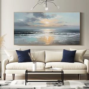 Op de afbeelding: Olieverfschilderij van een zeegezicht bij zonsondergang. Het kunstwerk toont een rustige oceaan met golven, een heldere zon en een bewolkte lucht. Het hangt boven een cr&egrave;mekleurige bank met marineblauwe kussens.