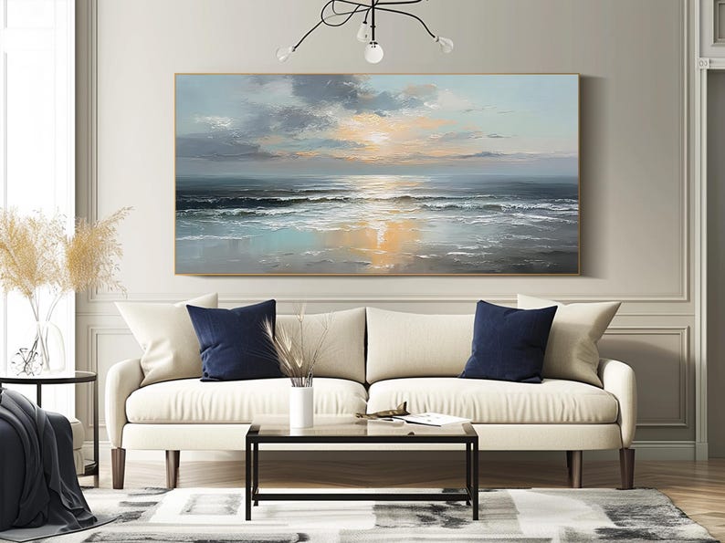 Op de afbeelding: Olieverfschilderij van een zeegezicht bij zonsondergang. Het kunstwerk toont een rustige oceaan met golven, een heldere zon en een bewolkte lucht. Het hangt boven een cr&egrave;mekleurige bank met marineblauwe kussens.
