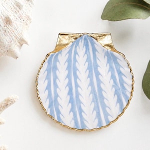 May include: A decorative seashell-shaped dish with a blue and white leaf pattern. The dish has a gold-colored rim and accents. The design is reminiscent of coastal decor.