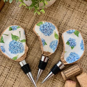 May include: Three blue and white hydrangea patterned bottle stoppers with silver tops. The stoppers are sitting on a woven brown surface.