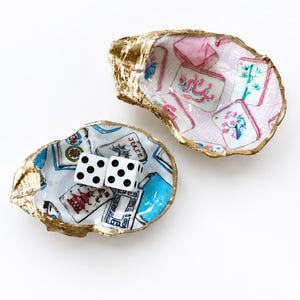 May include: Two decorative oyster shells with gold-painted edges. One shell features a pink and white pattern of mahjong tiles, while the other displays a blue and white pattern of mahjong tiles and includes two white six-sided dice with black pips.