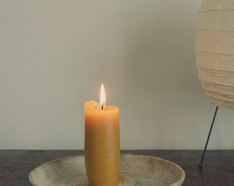 May include: A cylindrical golden yellow beeswax candle with a lit flame, resting in the center of a shallow, speckled beige ceramic dish. The candle has a small amount of melted wax pooling at its base on the textured surface of the plate.