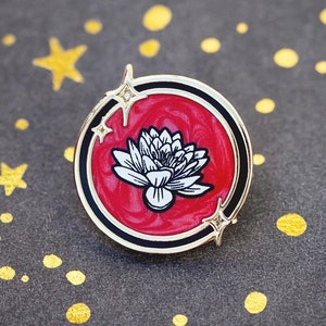 Puede incluir: Un pin de esmalte dorado con fondo rojo y un diseño de flor de loto blanca. El pin tiene un contorno negro y un detalle brillante.