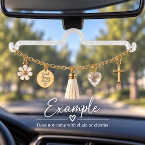 May include: A white acrylic car rearview mirror hanger with a gold-tone chain featuring five charms: a white daisy, a round gold disc with the text Good Things Ahead and a heart, a cream tassel, a clear heart-shaped crystal, and a gold cross.