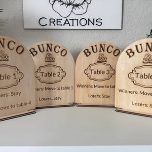 May include: Four wooden Bunco table signs. Each sign is arched at the top and features the word "BUNCO" above a decorative emblem and a table number. The signs also include instructions for winners and losers. The signs are made of light-colored wood.