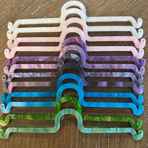 May include: A collection of ten acrylic hangers arranged in a vertical stack. Each hanger features a central arched hook and two small circular loops at the ends. The hangers display a variety of colors including white, pink, purple, blue, and green.