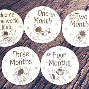 May include: Set of six wooden baby milestone discs with an astronaut theme. The discs feature a welcome sign with the name Elijah, and monthly milestones from one to six months. Each disc has a different astronaut design and stars.