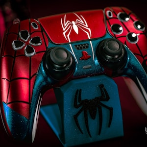 May include: A custom-painted Spider-Man-themed game controller. The controller features a red and blue color scheme with a white spider emblem on the top. The controller is resting on a blue stand with a black spider logo.