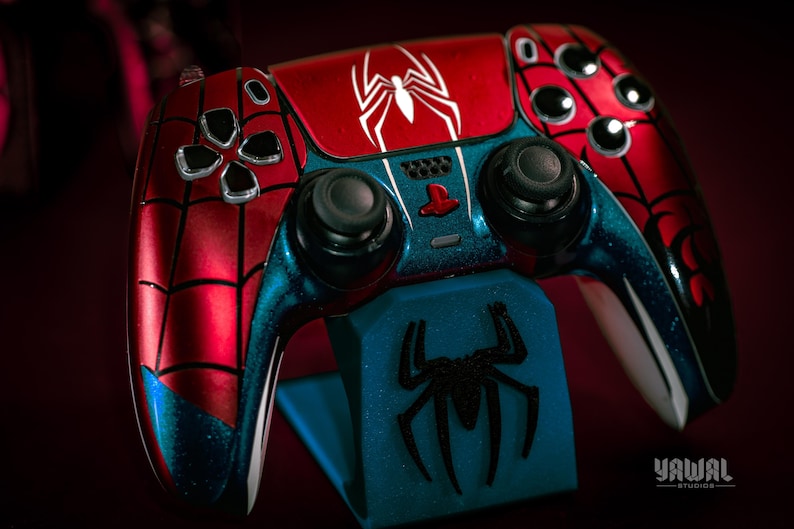 May include: A custom-painted Spider-Man-themed game controller. The controller features a red and blue color scheme with a white spider emblem on the top. The controller is resting on a blue stand with a black spider logo.