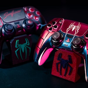 May include: Two custom-designed game controllers with matching stands. The controllers feature a red and black color scheme with spiderweb and lightning bolt designs. The stands have a spider logo. The word "Sunflower" is visible on one controller.