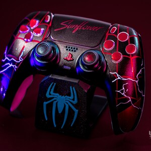 May include: Customised black and red video game controller with a matching stand. The controller has a spider-web design, red buttons, and the word "Sunflower". The stand displays a blue spider logo.