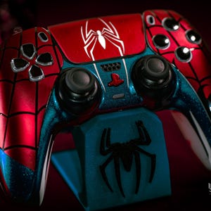 May include: A custom-painted video game controller in a Spider-Man theme. The controller features a red and blue color scheme with a white spider logo. The controller is on a blue stand with a black spider logo.