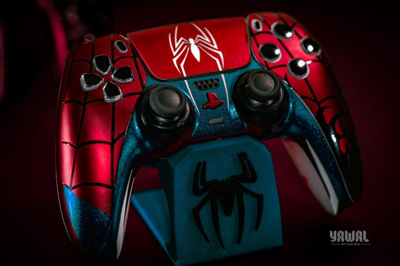 May include: A custom-painted video game controller in a Spider-Man theme. The controller features a red and blue color scheme with a white spider logo. The controller is on a blue stand with a black spider logo.
