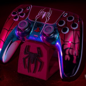 May include: A custom red and black Spider-Man-themed game controller with a matching stand. The controller features a white spider emblem and blue accents. The stand has a black spider logo. The controller is designed for gaming.