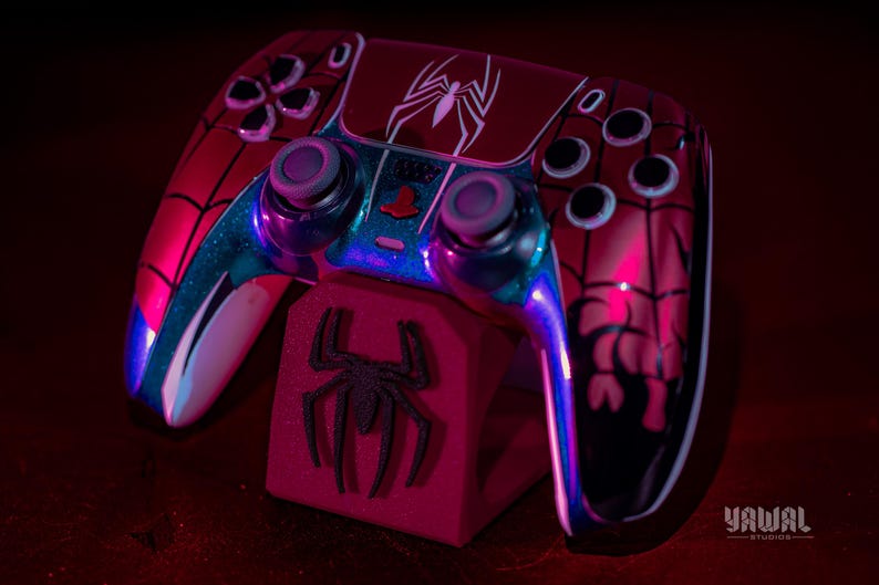 May include: A custom red and black Spider-Man-themed game controller with a matching stand. The controller features a white spider emblem and blue accents. The stand has a black spider logo. The controller is designed for gaming.