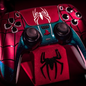 May include: A custom red and blue Spider-Man-themed video game controller. The controller features a white spider logo and web design. The controller is on a red stand with a black spider logo. The controller has black buttons.