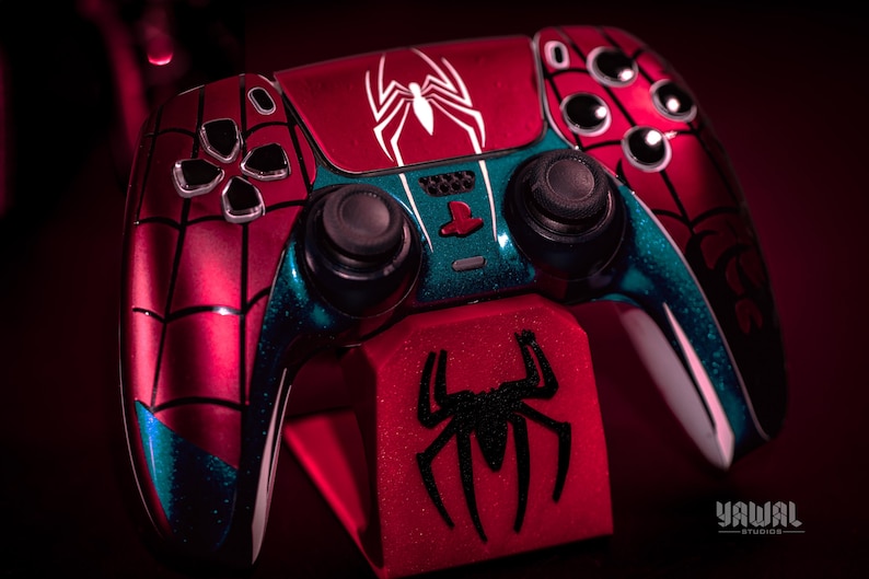 May include: A custom red and blue Spider-Man-themed video game controller. The controller features a white spider logo and web design. The controller is on a red stand with a black spider logo. The controller has black buttons.