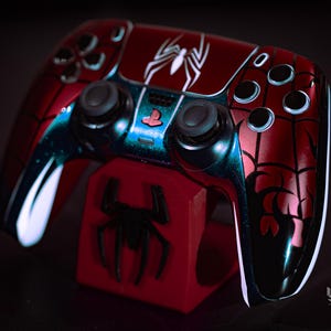 May include: A custom red and black Spider-Man-themed video game controller with a matching red stand. The controller features a white spider emblem and web detailing. The stand has a black spider emblem. The controller is for a PlayStation.
