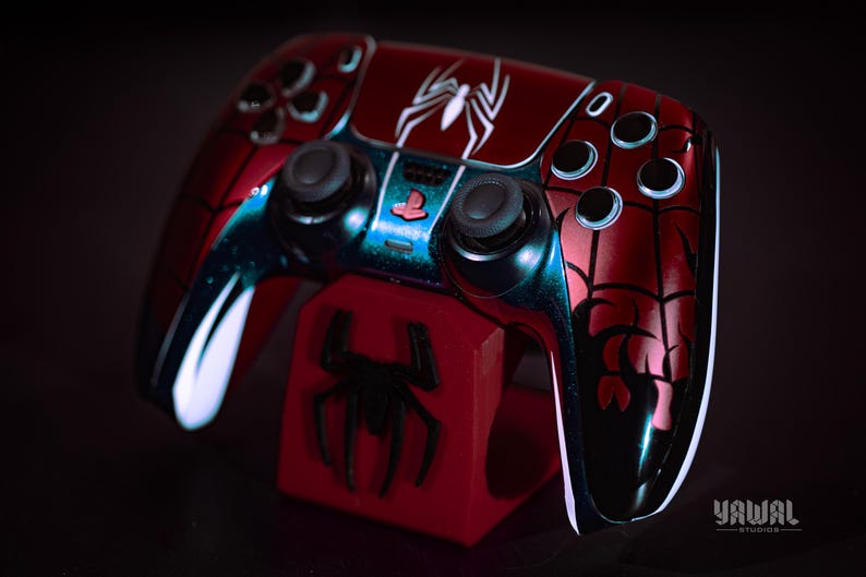 May include: A custom red and black Spider-Man-themed video game controller with a matching red stand. The controller features a white spider emblem and web detailing. The stand has a black spider emblem. The controller is for a PlayStation.
