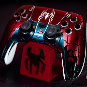 May include: A custom red and black Spider-Man-themed video game controller. The controller features a white spider emblem and web design. It rests on a red stand with a black spider logo. The controller has black buttons and joysticks.