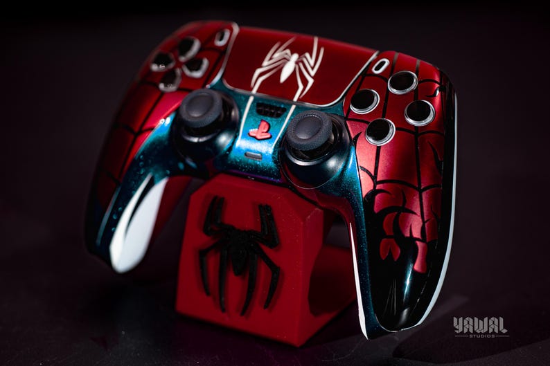 May include: A custom red and black Spider-Man-themed video game controller. The controller features a white spider emblem and web design. It rests on a red stand with a black spider logo. The controller has black buttons and joysticks.