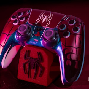 May include: A custom-painted video game controller with a Spider-Man theme. The controller is primarily red with black spiderweb detailing and a white spider emblem. The design includes blue and purple accents. A red stand with a spider logo supports the controller.